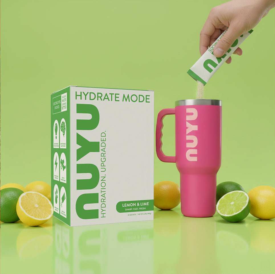 Hydrate Mode lemon lime electrolyte drink mix being poured into pink Nuyu tumbler with fresh citrus fruits and product box on green background