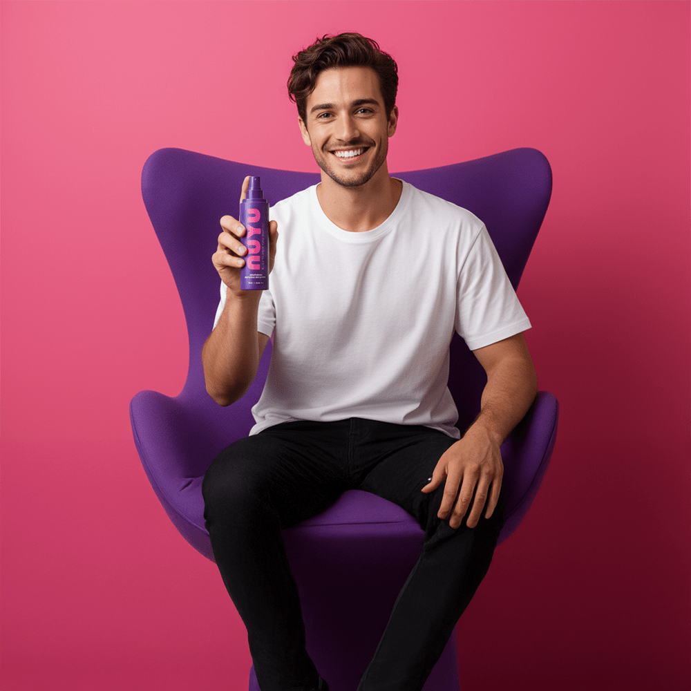 Smiling man sitting on purple chair holding Muscle & Joint Magnesium NUYU spray bottle against pink background